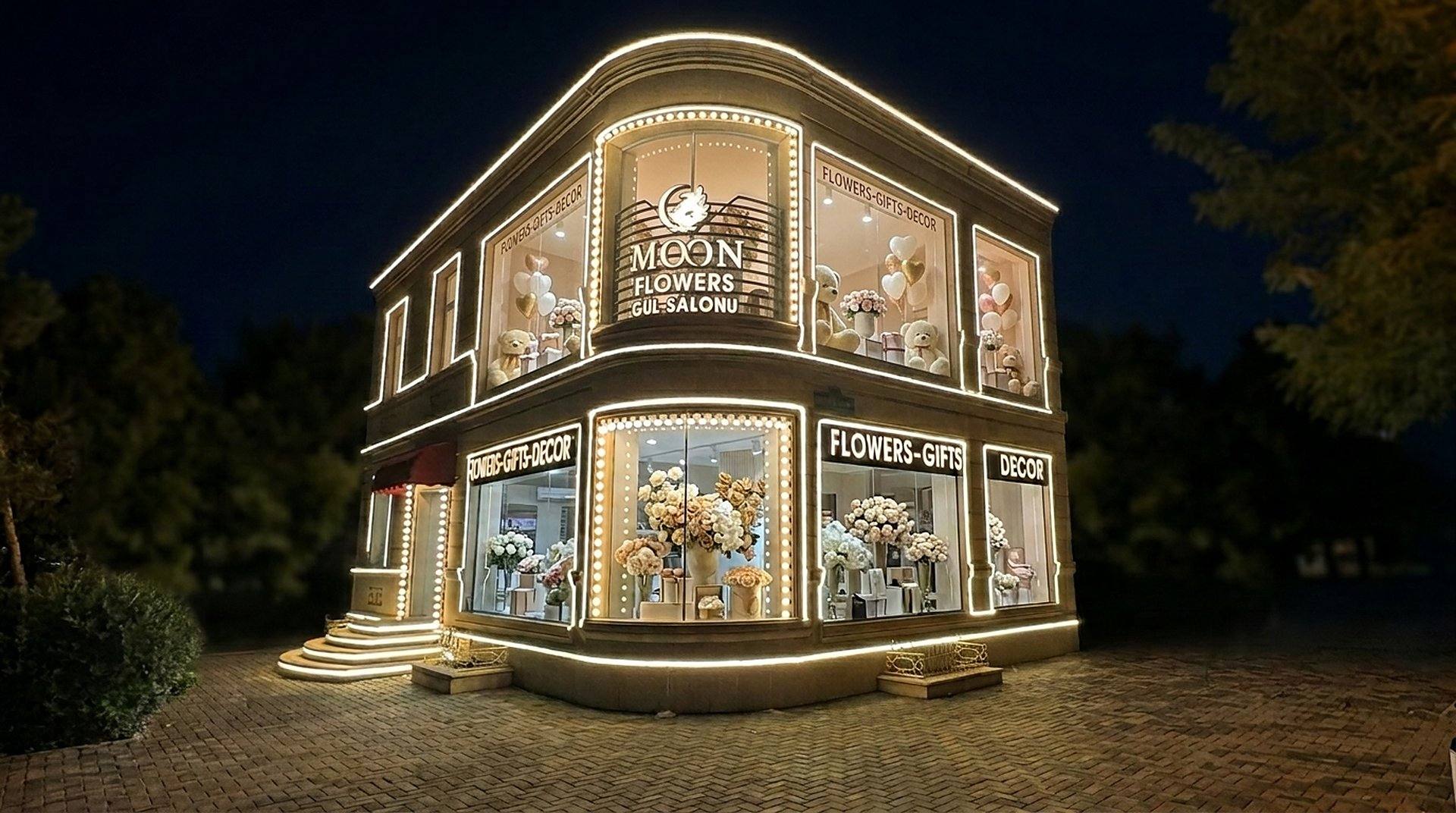 Moon Flowers gül salonunun gecə fasadı: qızılı işıqlı vitrinlər və buketlər